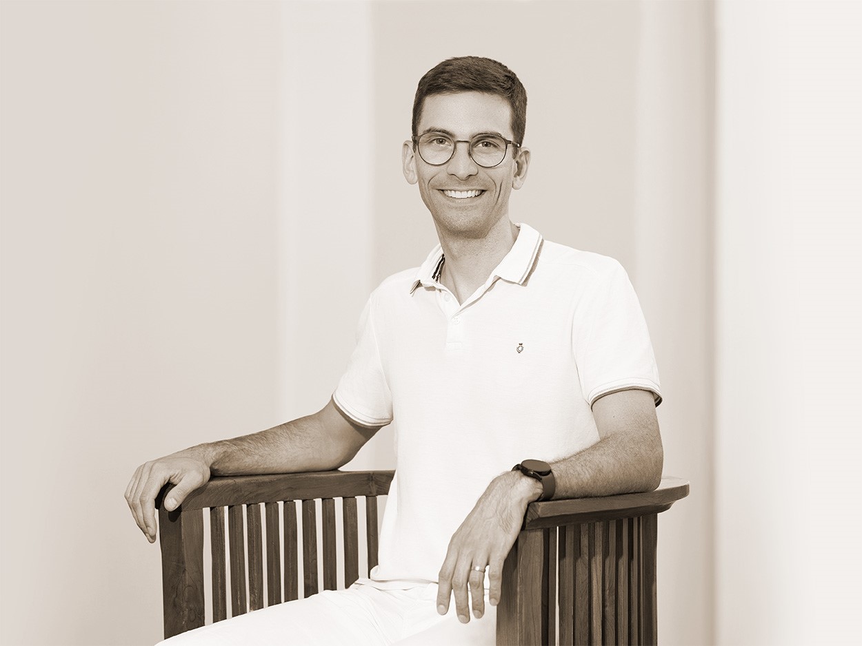 Raphael Baptista Gallo ist weiß gekleidet, trägt eine runde Brille, sitzt auf einem Stuhl und lächelt.